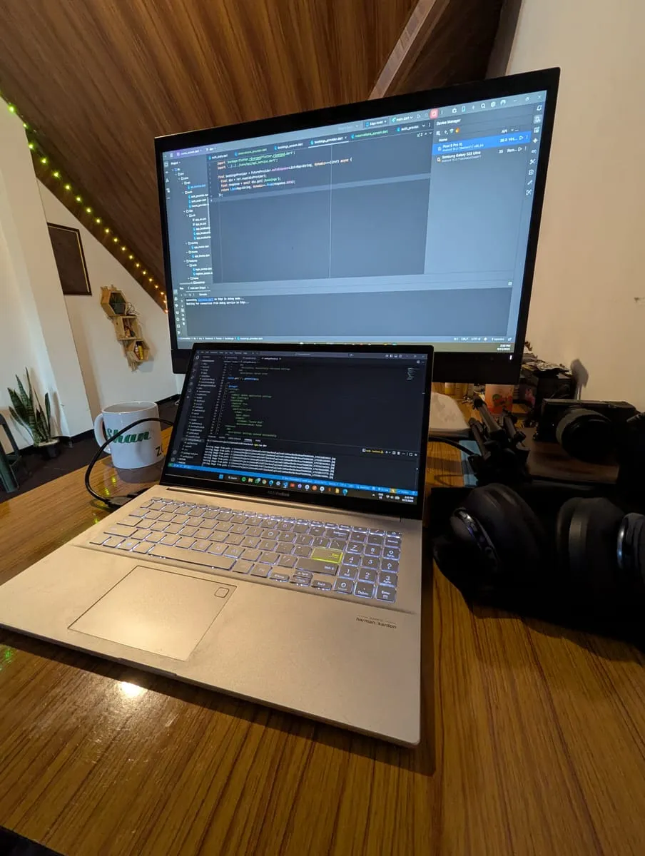 A laptop and monitor on a wooden desk lit by fairy lights, with a mug and a plant beside them.