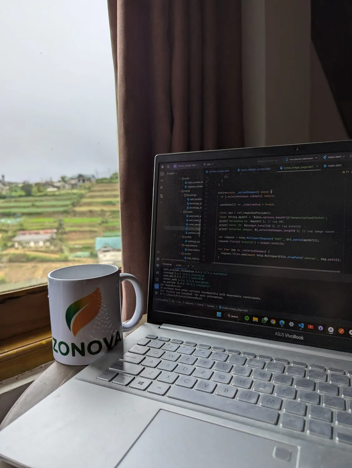 A branded Zonova mug on a studio windowsill beside a laptop showing Dart and Flutter source code, with the misty terraced hills of Nuwara Eliya through the glass.