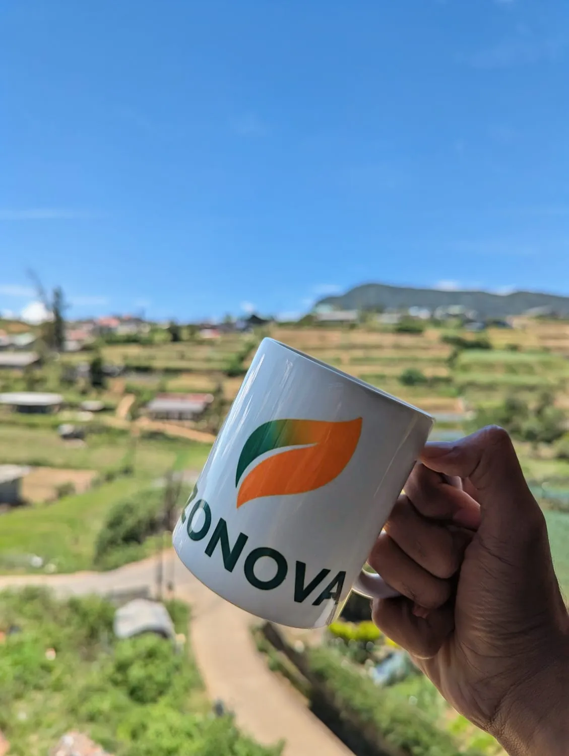A Zonova Tech mug held up against the terraced hillsides and village rooftops of Nuwara Eliya.