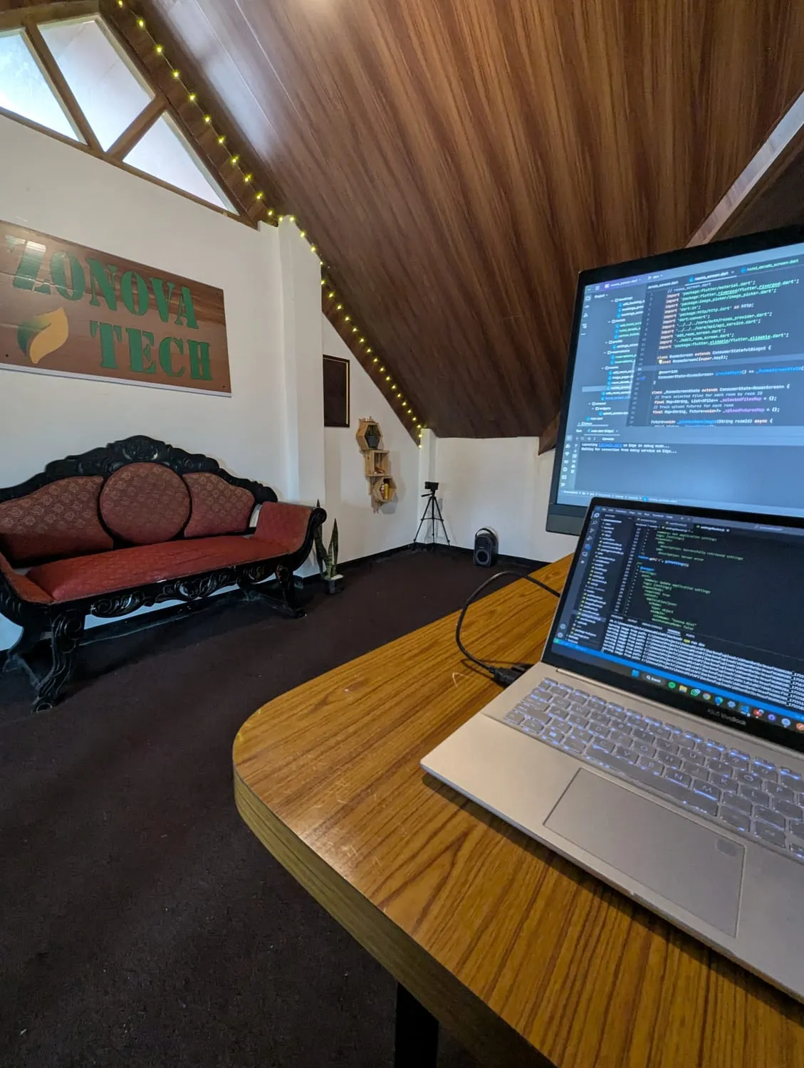 A desk in the Zonova Tech studio: a laptop and an external monitor showing code, looking across the room to the studio's hand-painted wooden sign.